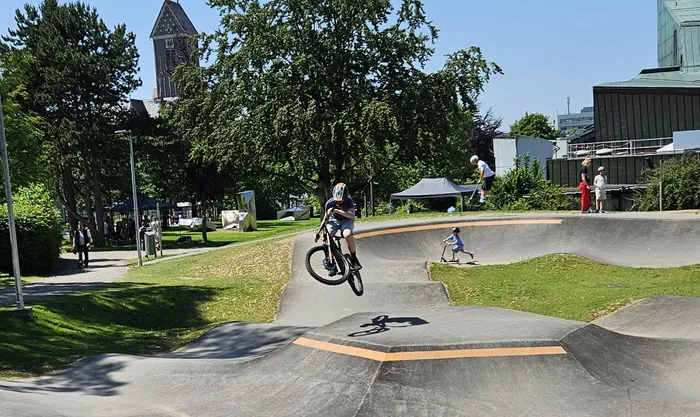 Ein Junge fährt mit dem Fahrrad in der Luft über einer Rampe in einem Skatepark. Im Hintergrund sind weitere Personen und Gebäude zu sehen.