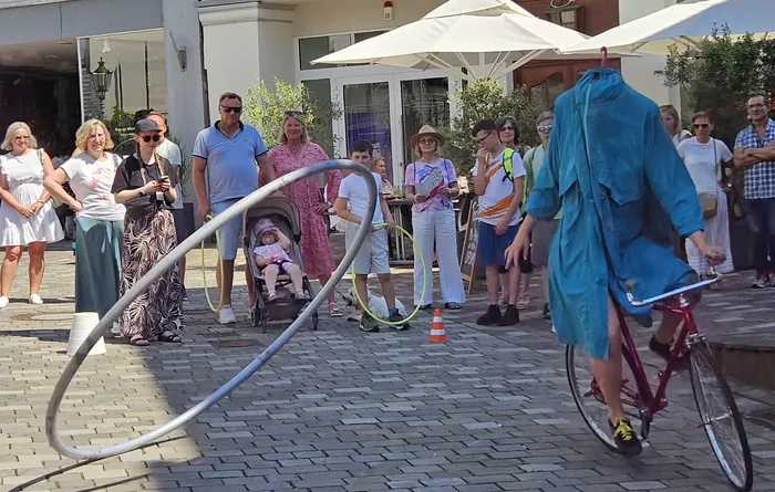 Ein Mann fährt auf einem roten Hochrad und führt mit einem Reifen einen Kreis vor einer Zuschauermenge auf einem Platz auf. Einige Zuschauer stehen, andere sitzen an Tischen unter Sonnenschirmen.