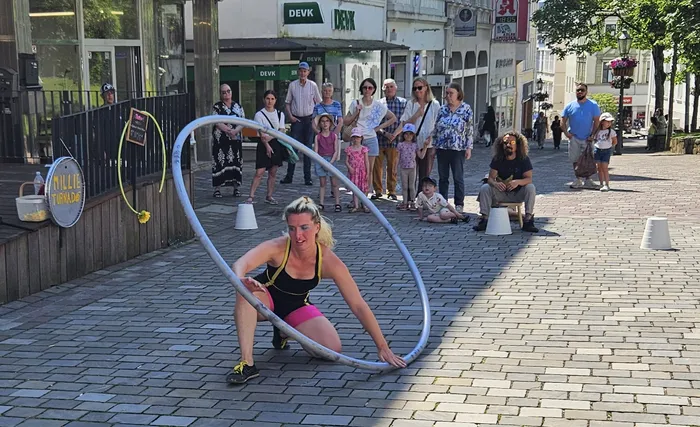 Eine Frau in Sportkleidung führt eine Übung mit einem großen Reifen auf einem gepflasterten Platz vor Zuschauern auf. Im Hintergrund sind Geschäfte und Passanten zu sehen.