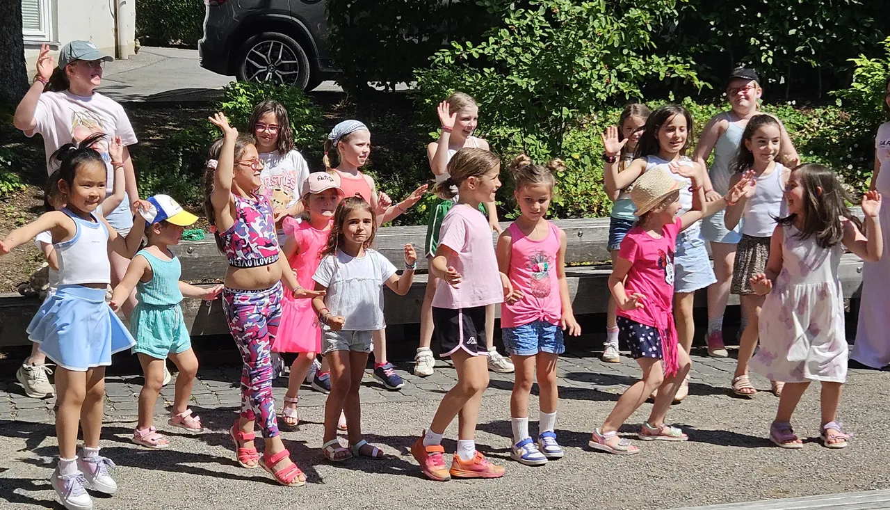 Eine Gruppe von Kindern in farbenfroher Kleidung tanzt und gestikuliert auf einer Straße. Im Hintergrund sind weitere Kinder und ein Auto zu sehen.