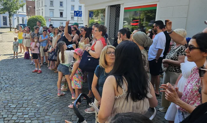Menschen stehen und sitzen an einer Pflasterstraße und beobachten eine Tanzaufführung. Einige applaudieren, andere filmen mit ihren Handys. Im Hintergrund sind Häuserfassaden und ein rotes Auto zu sehen.
