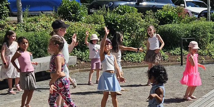 Eine Gruppe Kinder tanzt im Freien auf einem Platz. Einige heben die Arme, andere bewegen sich im Rhythmus der Musik. Im Hintergrund sind Büsche und parkende Autos zu sehen.