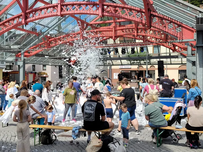Menschen und Kinder stehen inmitten von aufspritzendem weißem Schaum unter einer großen roten Metallkonstruktion.