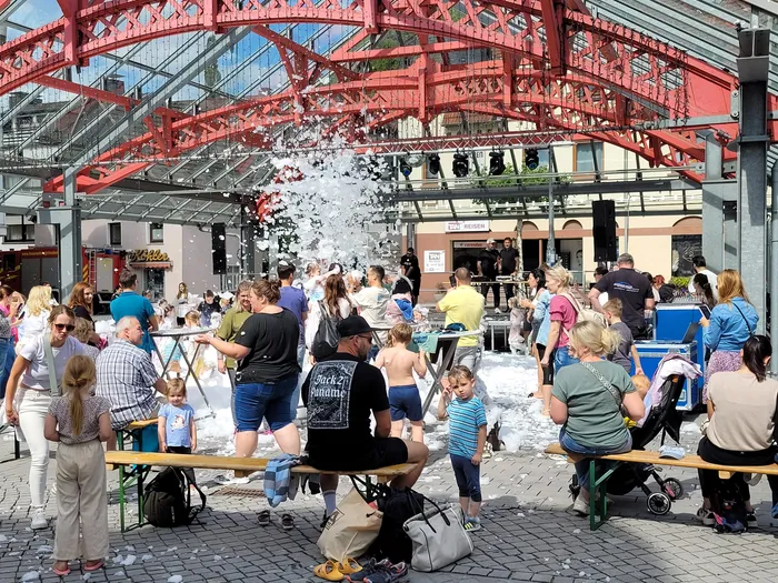Menschen und Kinder spielen bei einer Schaumparty unter einer großen roten Metallkonstruktion auf dem Alten Markt.