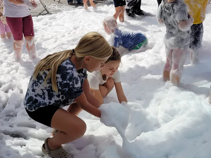 Ein Mädchen spielt im Vordergrund mit weißem Schaum, während andere Kinder im Hintergrund in der Schaumlandschaft stehen.