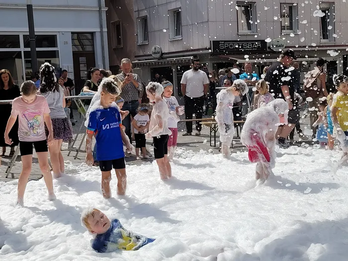 Kinder spielen in einer dicken Schicht aus weißem Schaum auf dem Alten Markt, während im Hintergrund Erwachsene zusehen.