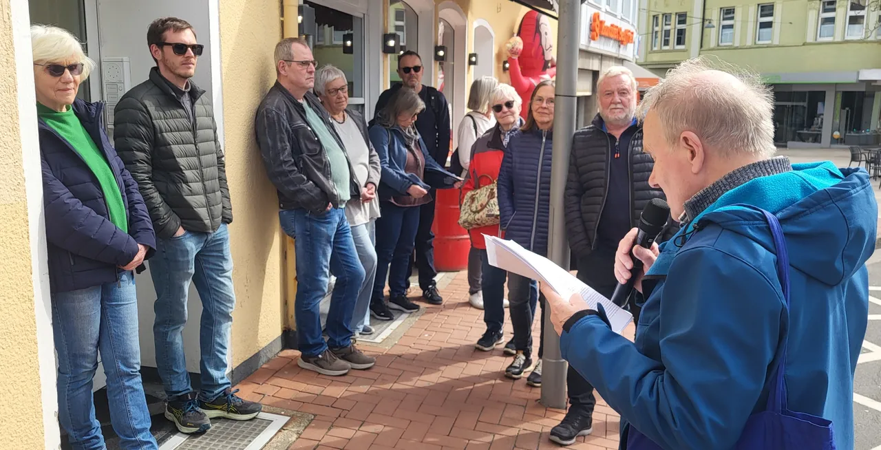 Mann mit Mikrofon spricht vor einer Gruppe von Menschen auf einem sonnigen Platz, während er Papiere hält. Im Hintergrund sind Geschäfte und ein rotes Banner zu sehen.