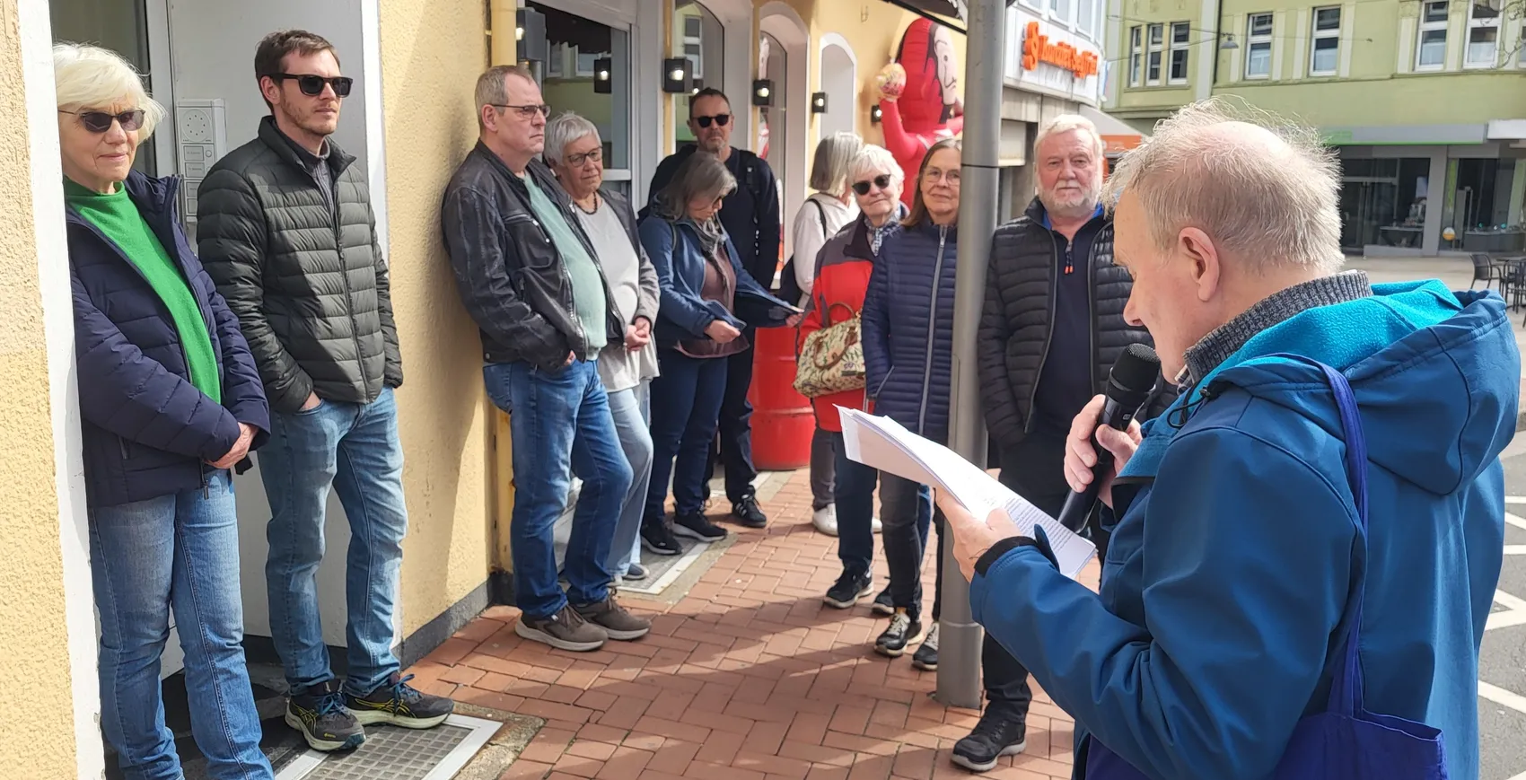 Mann mit Mikrofon spricht vor einer Gruppe von Menschen auf einem sonnigen Platz, während er Papiere hält. Im Hintergrund sind Geschäfte und ein rotes Banner zu sehen.