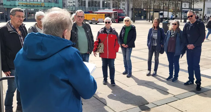 Eine Gruppe von etwa zehn Personen steht auf dem Rathausplatz zusammen und lauscht einer Führung. Im Hintergrund ist eine Bank und ein gelber LKW zu sehen.