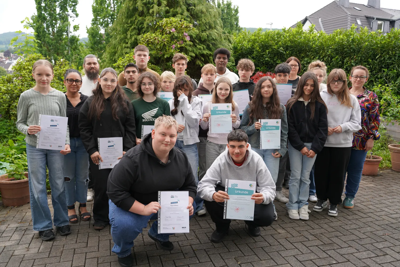 Gruppe von Schülerinnen und Schülern der Zeppelinschule hält Urkunden in den Händen, umgeben von Lehrkräften im Schulhof. Zwei Schüler knien in der ersten Reihe und halten ihre Urkunden hoch.