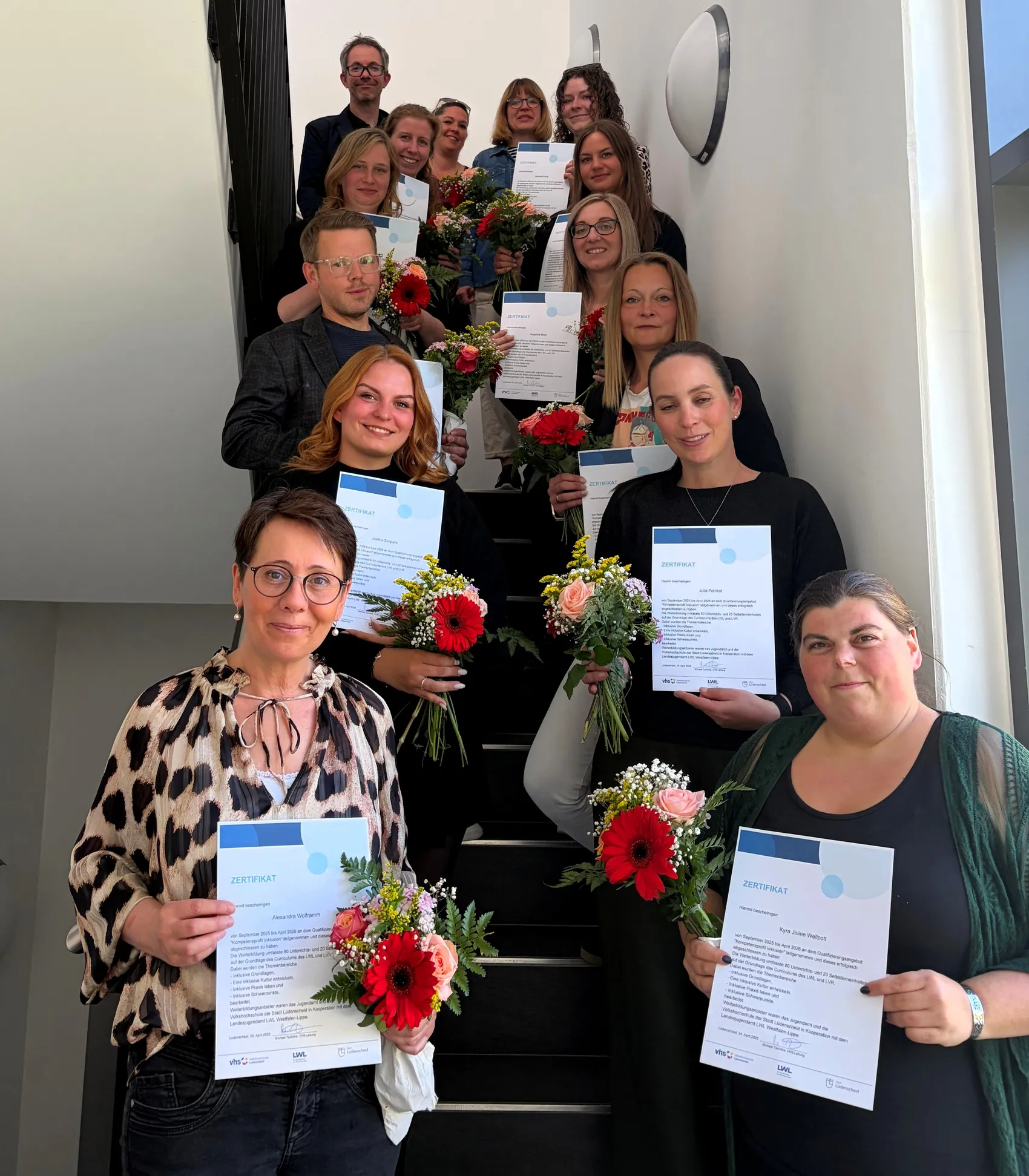 Eine Gruppe von 14 Fachkräften der Lüdenscheider Kindertageseinrichtungen steht auf einer Treppe und hält Zertifikate und Blumensträuße. Sie haben eine Fortbildung zum Thema Inklusion abgeschlossen.
