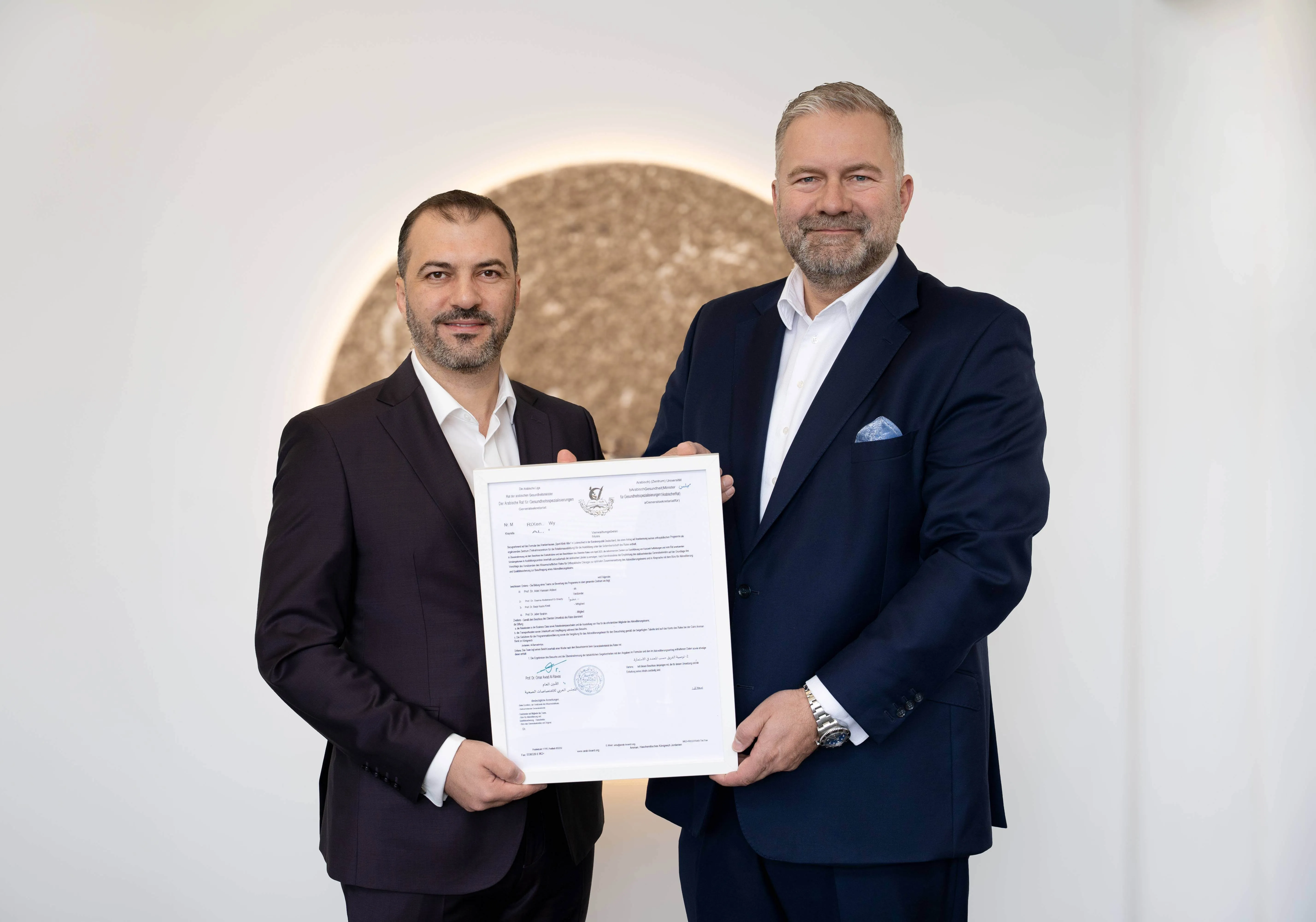 Zwei Männer in Anzügen halten ein Zertifikat. Links Prof. Mohamed Elashhab, rechts Dirk Burghaus, beide lächeln in die Kamera.