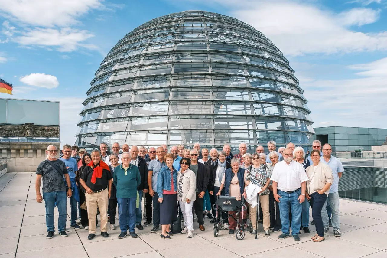 Eine Gruppe von etwa 40 Personen steht vor dem Reichstagsgebäude in Berlin für ein Gruppenfoto zusammen. Im Hintergrund ist die gläserne Kuppel des Reichstags bei blauem Himmel zu sehen.