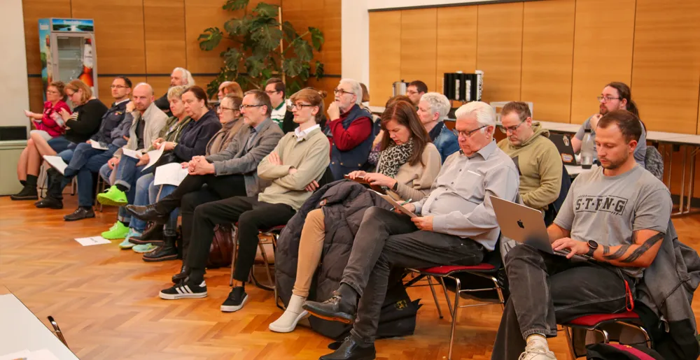 Zuschauer sitzen auf Stühlen in einem hellen Raum und verfolgen eine Ratssitzung. Einige nutzen Laptops oder machen sich Notizen.
