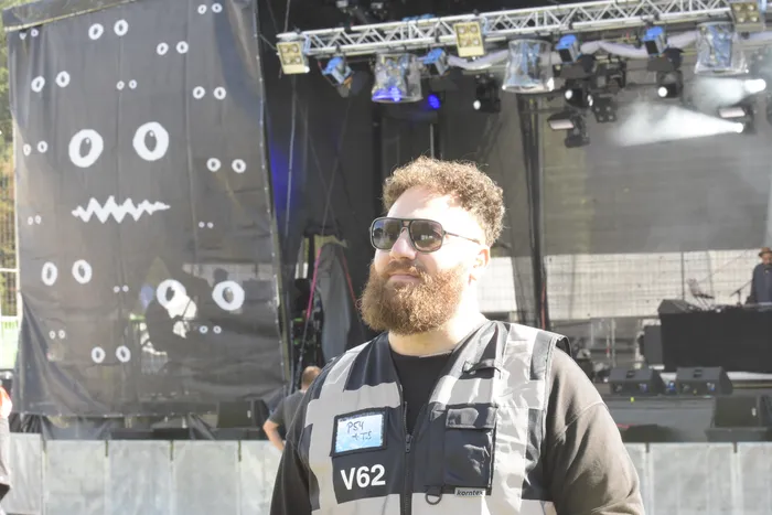 Ein bärtiger Mann mit Sonnenbrille und Weste vor einer Bühne mit einem schwarzen Hintergrund mit Augen- und Mundmotiven.
