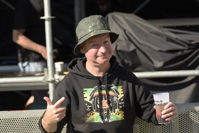 Shorty mit grünem Bucket Hat und schwarzem Hoodie macht eine Handgeste und hält einen Becher.