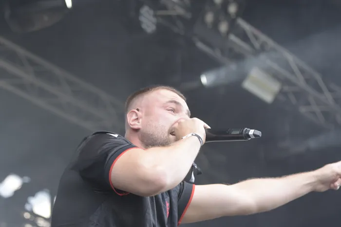 Ein bärtiger Mann in einem schwarzen T-Shirt singt in ein Mikrofon auf einer Bühne vor Bühnenlicht und Traversen.