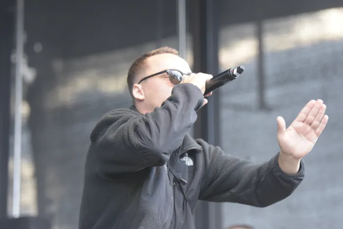 Ein Mann mit Sonnenbrille und schwarzer Jacke singt in ein Mikrofon und hält seine andere Hand erhoben.