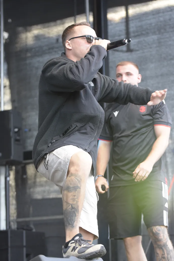 Ein Mann mit Sonnenbrille und schwarzer Fleecejacke singt in ein Mikrofon. Er trägt beige Shorts und hat ein großes Tattoo am Oberschenkel.