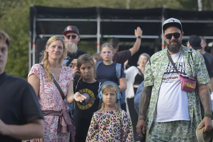 Eine Gruppe von Menschen vor einer Bühne, darunter eine Frau in einem gemusterten Kleid, ein Kind mit blauen Kopfhörern und ein Mann in einem grün gemusterten Outfit.