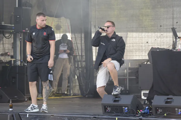 Zwei Männer performen auf einer Bühne; einer trägt ein schwarzes T-Shirt, der andere eine schwarze Jacke, Sonnenbrille und helle Shorts.