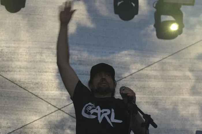 Ein Mann mit schwarzer Kappe und T-Shirt mit der Aufschrift „CARL“ hält ein Mikrofon und hebt einen Arm.