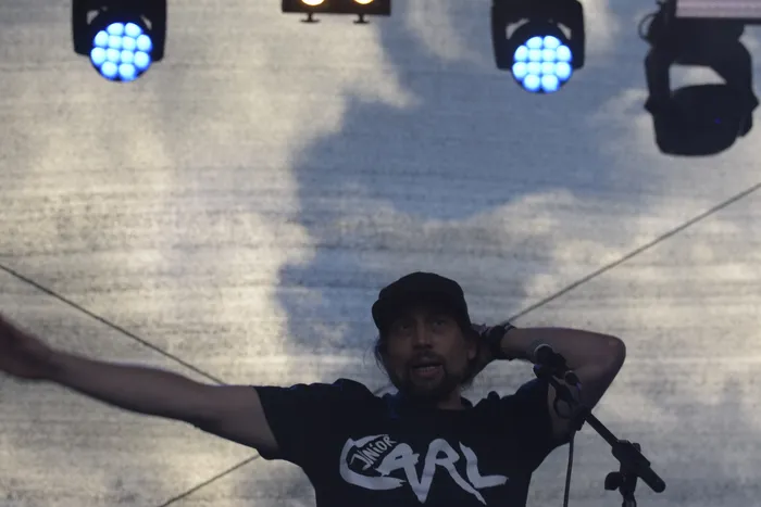 Ein Mann mit Cap und schwarzem T-Shirt mit der Aufschrift „JUNIOR CARL“ steht mit ausgebreiteten Armen auf einer Bühne vor einem bewölkten Himmel.