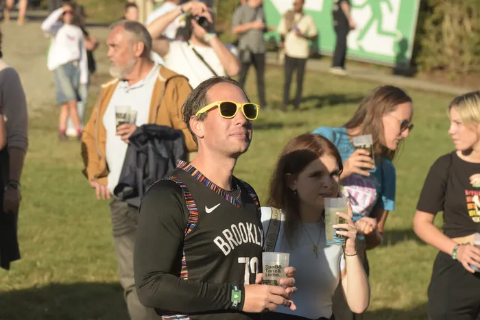 Ein Mann mit gelber Sonnenbrille und einem schwarzen Brooklyn-Trikot blickt nach oben, neben ihm eine Frau mit einem Getränkebecher.