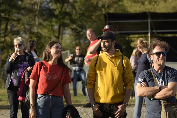 Menschen stehen vor einer Bühne bei einem Open-Air-Event, darunter eine Frau in einem roten T-Shirt und ein Mann in einem gelben Kapuzenpullover.