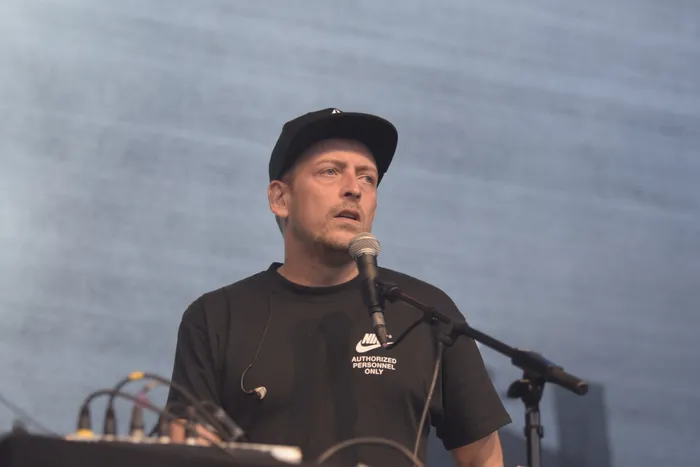 Hexer mit schwarzer Kappe und einem schwarzen T-Shirt mit der Aufschrift „Nike Authorized Personnel Only“ steht vor einem Mikrofon.
