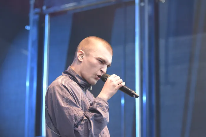 Ein Mann mit kurzen Haaren und grauer Jacke singt in ein Mikrofon auf einer blau beleuchteten Bühne.