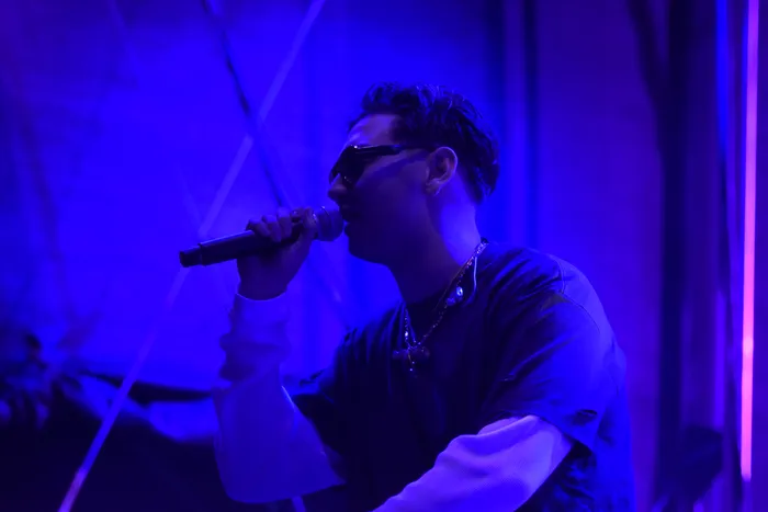Sampagne singt im Profil in ein Mikrofon unter blauem und violettem Bühnenlicht.