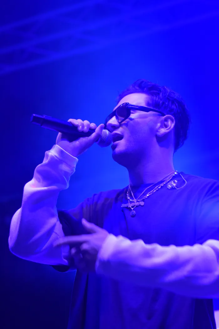 Sampagne singt unter blauem Bühnenlicht in ein Mikrofon und trägt eine Sonnenbrille sowie Halsketten.