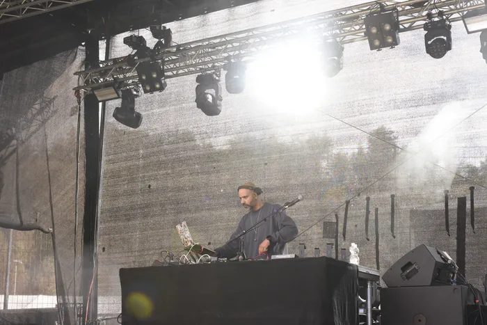 Ein Mann mit Kappe bedient DJ-Equipment an einem Pult auf einer Bühne unter hellen Scheinwerfern.