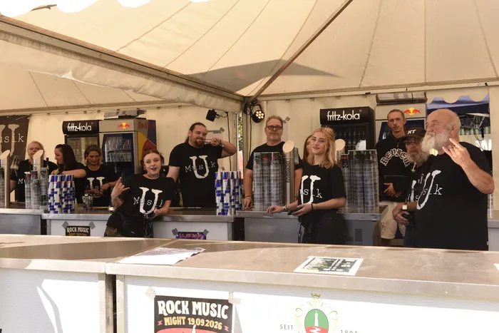 Mehrere Personen in schwarzen T-Shirts mit der Aufschrift „MUSIK“ stehen hinter einem Tresen unter einem Festzelt bei der „Rock Music Night“.