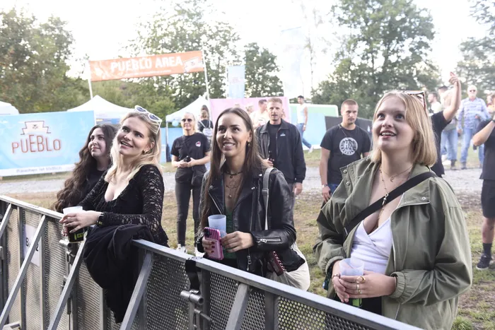 Vier junge Frauen stehen lächelnd hinter einer Metallabsperrung bei einem Festivalgelände mit Zelten und dem Schriftzug „PUEBLO AREA“ im Hintergrund.