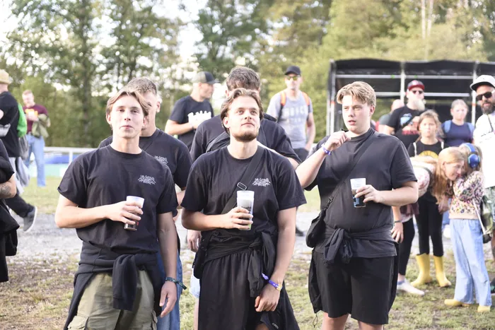 Drei junge Männer in schwarzen T-Shirts halten Getränke in Plastikbechern vor einer Bühne und einer Menschenmenge im Freien.