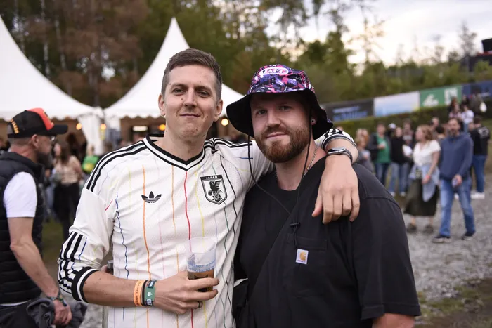 Zwei Männer posieren eng umschlungen vor Zelten und einer Menschenmenge; einer trägt ein weißes Japan-Fußballtrikot und hält ein Bierglas.