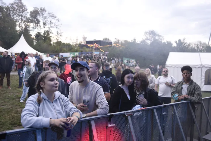 Eine Gruppe junger Menschen steht hinter einer Metallabsperrung bei einem Open-Air-Event und blickt in Richtung der Bühne.