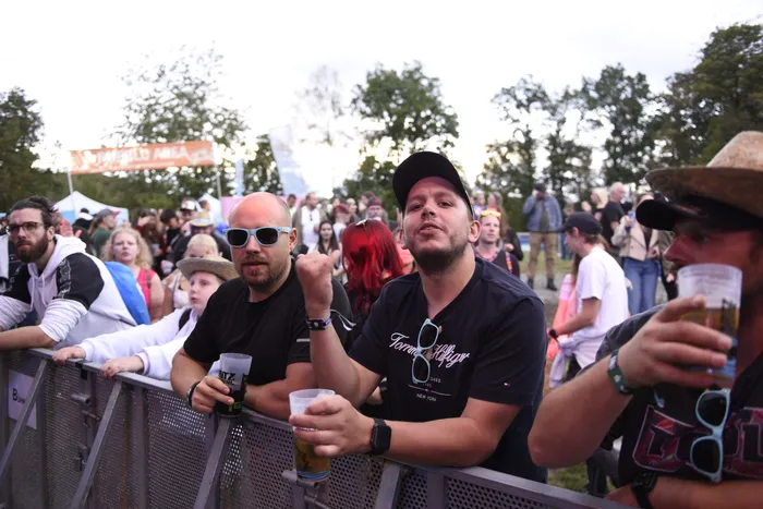 Menschen stehen an einer Metallabsperrung und halten Getränke bei einem Open-Air-Event.
