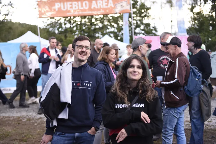 Ein lächelnder Mann mit Brille und eine Frau stehen in einer Menschenmenge vor einem Banner mit der Aufschrift „PUEBLO AREA“ bei einer Open-Air-Veranstaltung.