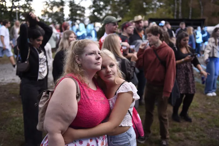 Zwei junge Frauen umarmen sich und blicken nach oben inmitten einer Menschenmenge bei einem Open-Air-Event.