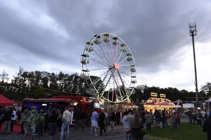 Ein beleuchtetes Riesenrad über einer Menschenmenge bei einem Open-Air-Festival unter bewölktem Abendhimmel.