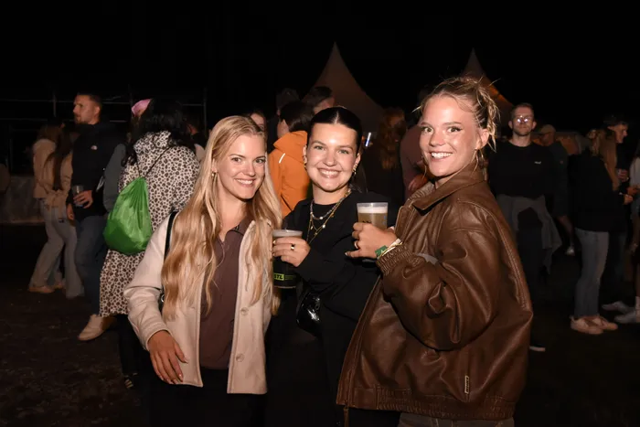 Drei lächelnde junge Frauen halten Getränke in den Händen und posieren bei einer nächtlichen Veranstaltung im Freien.