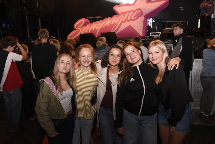 Fünf junge Frauen posieren lächelnd vor einer Bühne mit einem großen, pinkfarbenen Stern-Schild und dem Schriftzug „Sampagne“.