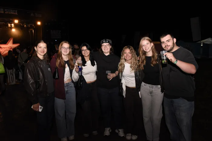 Eine Gruppe von sieben jungen Menschen posiert lächelnd mit Getränken vor einer beleuchteten Festivalbühne bei Nacht.