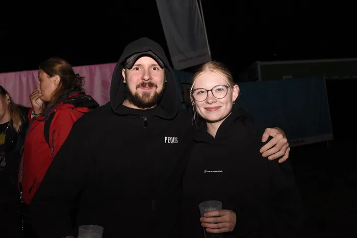 Ein lächelndes Paar in schwarzen Kapuzenpullovern hält Plastikbecher bei einer Abendveranstaltung.