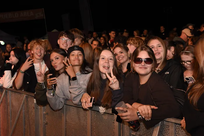Eine Gruppe junger Menschen lächelt und macht Handzeichen hinter einer Metallabsperrung bei einem Musikfestival.
