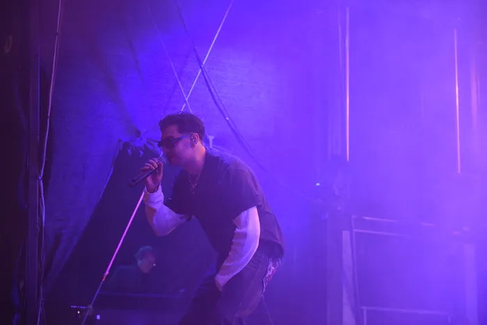 Ein Sänger mit Sonnenbrille singt unter violettem Bühnenlicht in ein Mikrofon.