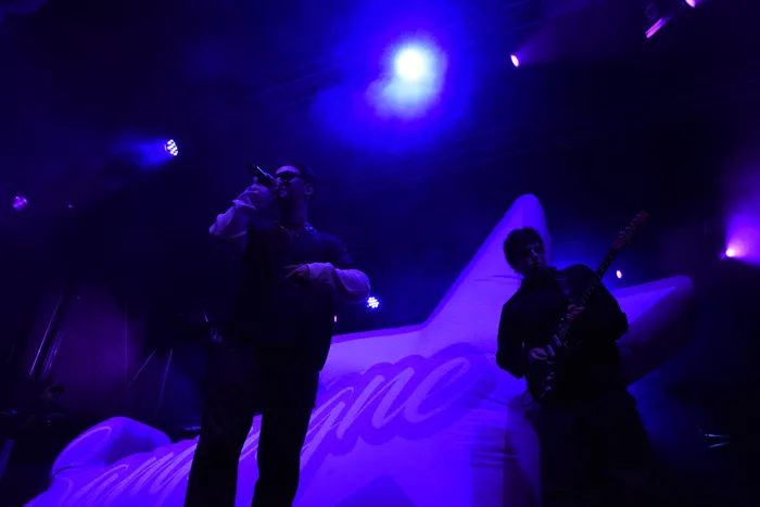 Sänger Sampagne performt mit einem Mikrofon auf einer Bühne unter blauem und violettem Licht neben einem Gitarristen.